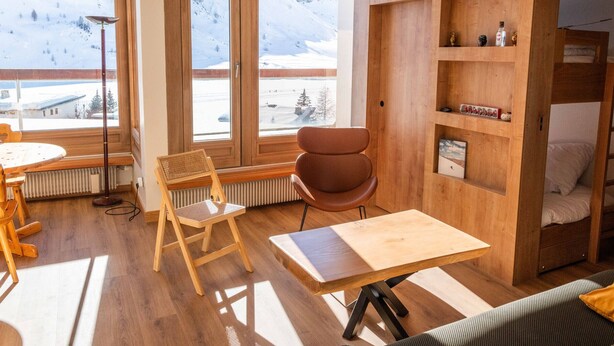 A modern room with wooden floors, a dining table, a chair, a small table, a radiator, a shelf with items, and a window with a snowy mountain view.