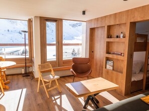 A modern room with wooden floors, a dining table, a chair, a small table, a radiator, a shelf with items, and a window with a snowy mountain view.