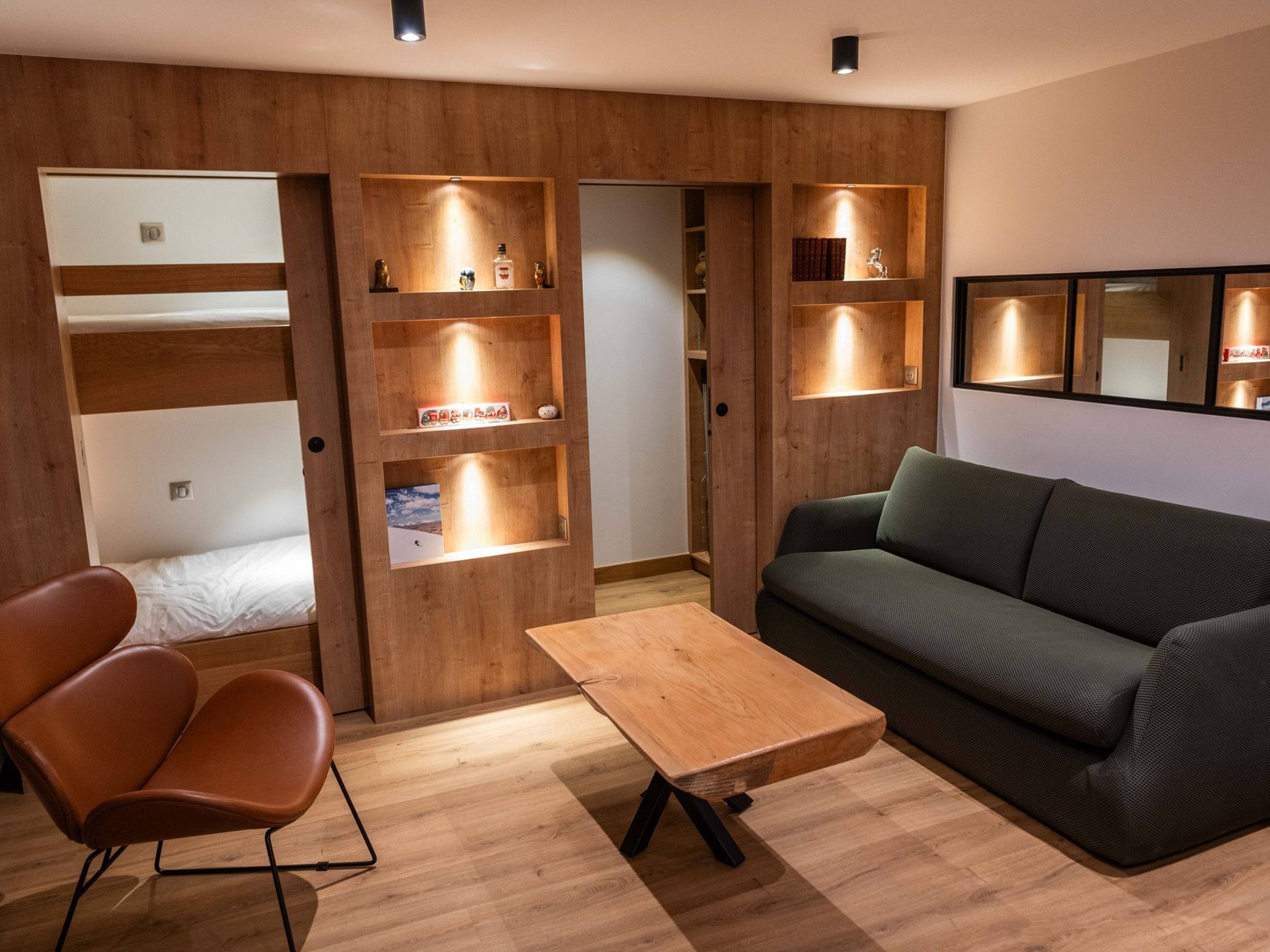 Une pièce moderne comprenant un canapé, une table en bois, une chaise et des étagères intégrées où sont disposés des livres et des objets décoratifs.