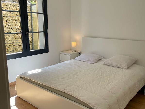 A bedroom with a bed, a nightstand, a lamp, and a window with a stone wall view.