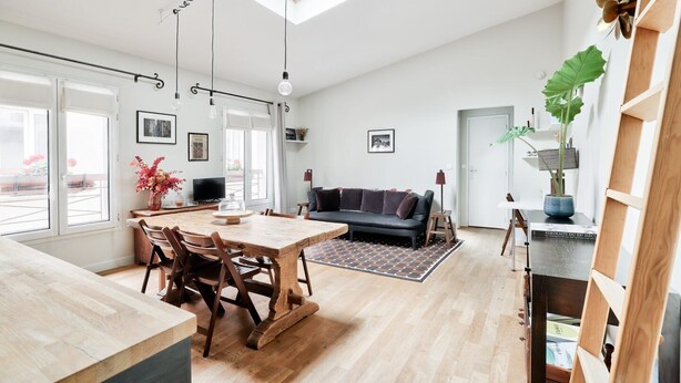 Amplia sala de estar con una mesa de comedor de madera, un sofá oscuro y un ventanal que proporciona luz natural.