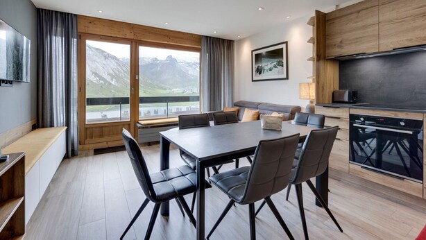 Un comedor moderno con una mesa grande y sillas, una cocina con armarios oscuros y una vista panorámica de montañas a través de amplios ventanales.