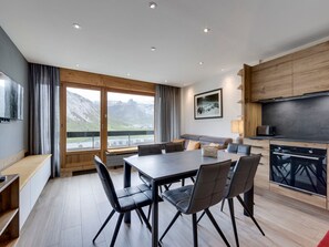 A modern dining area with a large table and chairs, a kitchen with dark cabinets, and a scenic mountain view through large windows.