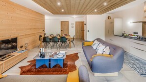 Ein modernes Wohnzimmer mit Holzbalkendecke, einem Essbereich mit Tisch für vier Personen, einer Couch mit Kissen, einem Flachbildfernseher und einer Küchenzeile mit Arbeitsfläche und Spüle.