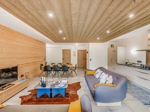 Ein modernes Wohnzimmer mit Holzbalkendecke, einem Essbereich mit Tisch für vier Personen, einer Couch mit Kissen, einem Flachbildfernseher und einer Küchenzeile mit Arbeitsfläche und Spüle.