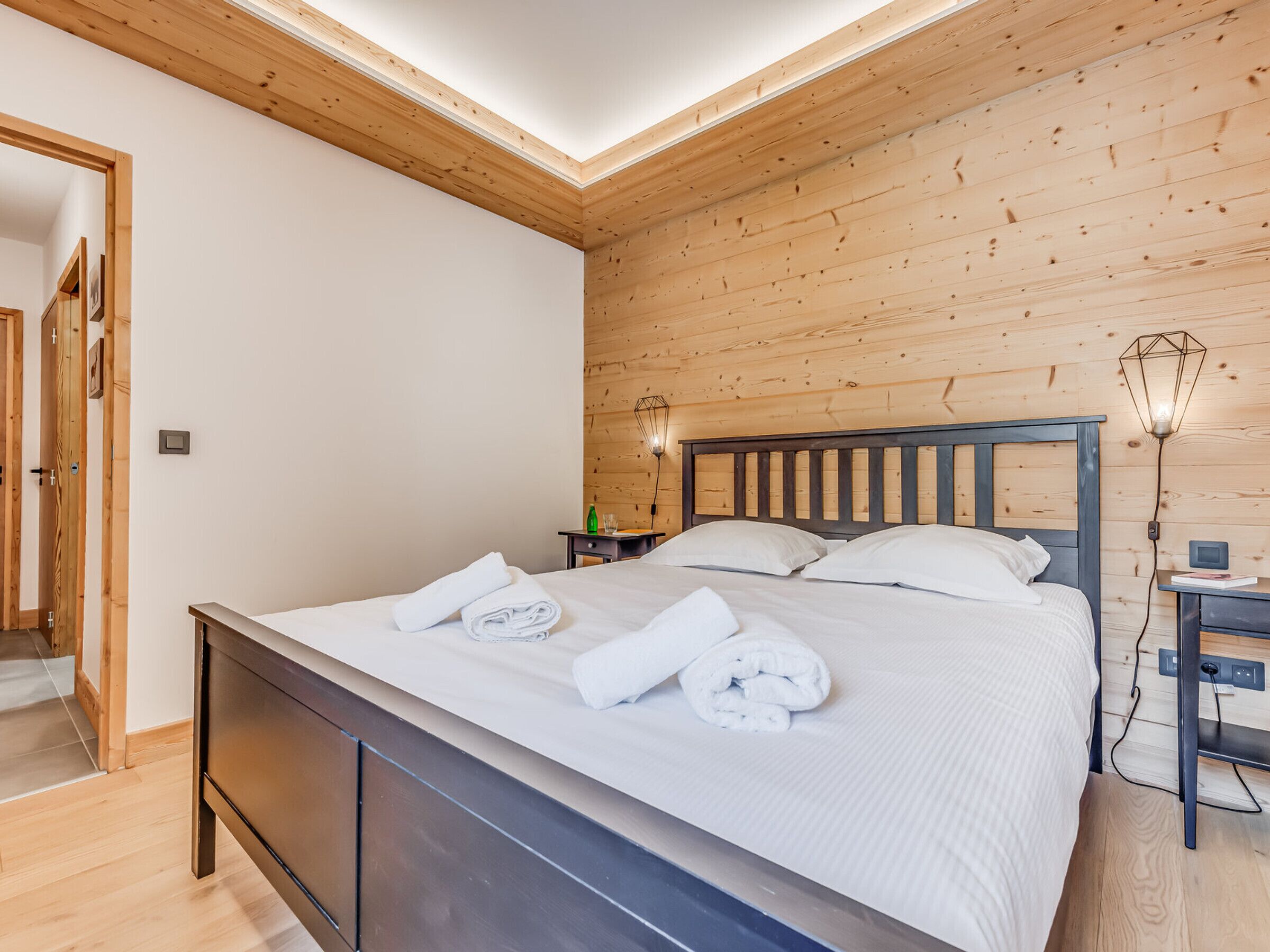 A bedroom with a wooden ceiling, a bed with white linens, a nightstand with a lamp, and a wooden door.