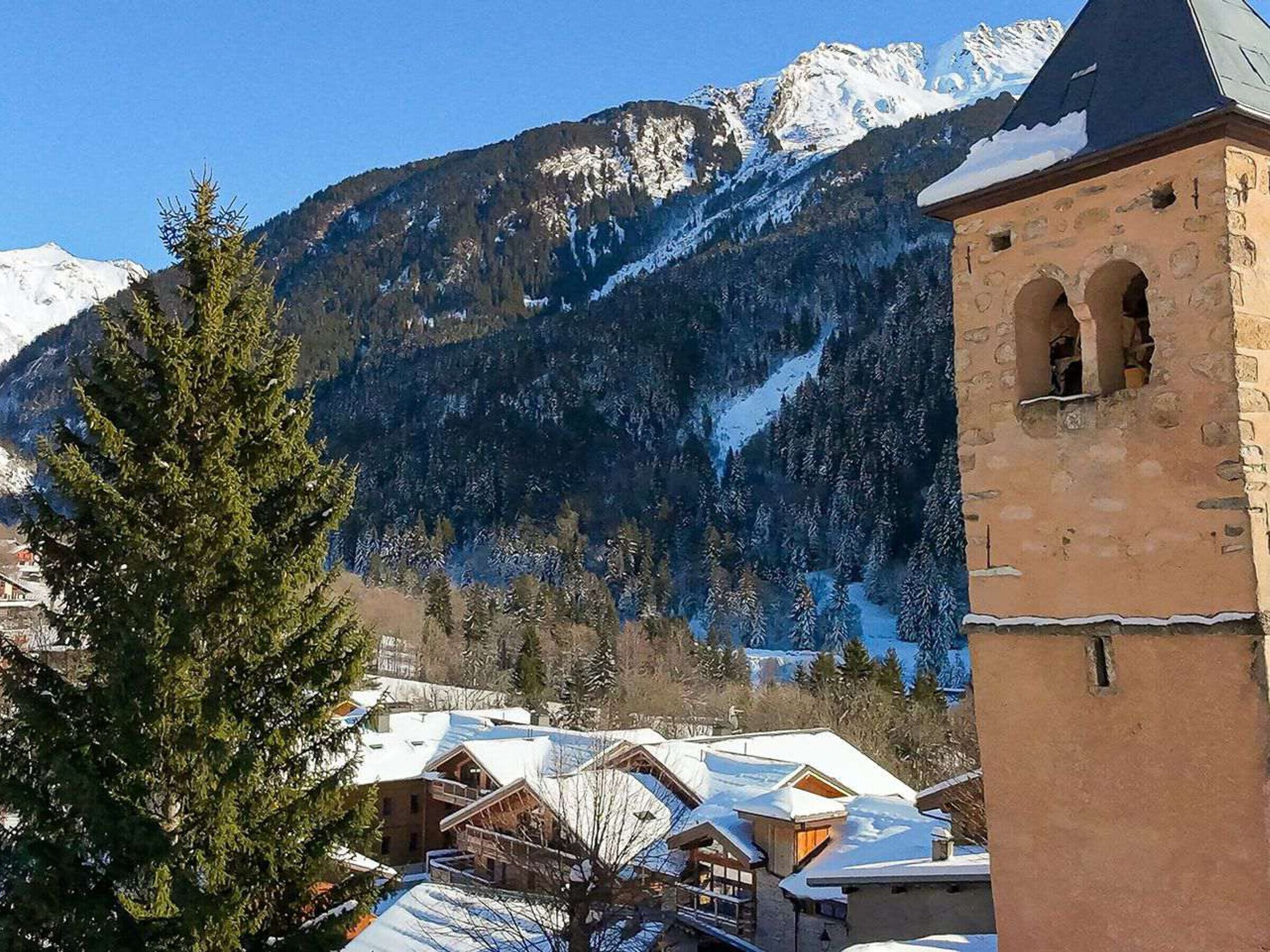 Un village de montagne aux toits enneigés, une église aux fenêtres en arc de cercle et un paysage boisé en arrière-plan.