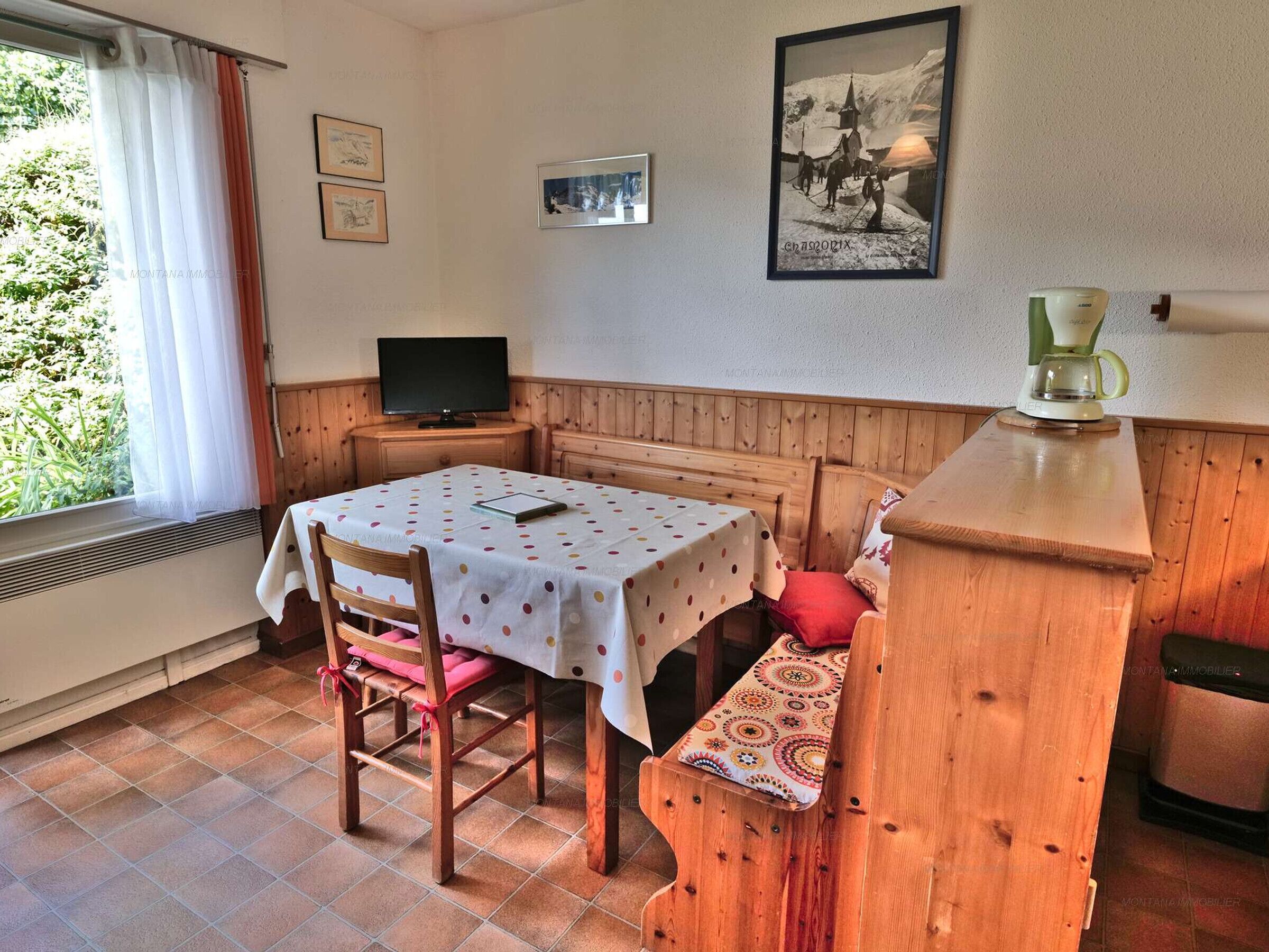 Une pièce chaleureuse avec un banc en bois, une table recouverte d’une nappe à pois, un téléviseur posé sur un meuble en bois et un tableau encadré accroché au mur.