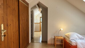 Un dormitorio con un armario de madera, una cama con almohada blanca, una mesita de noche de madera con una lámpara y vistas a una escalera que conduce a otra habitación.