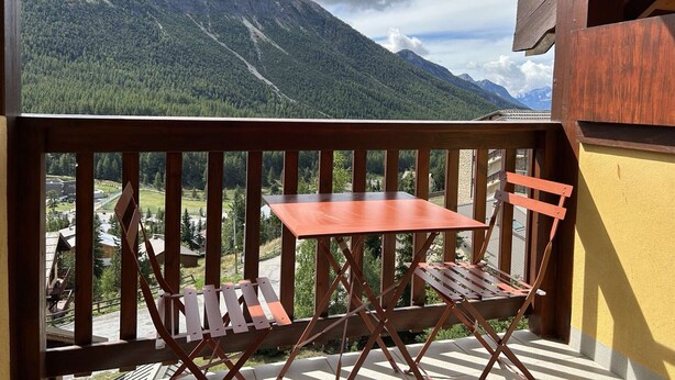 Un balcón con barandilla de madera, una mesa pequeña y dos sillas. Al fondo, se observa una cadena montañosa y un edificio con una pared amarilla.