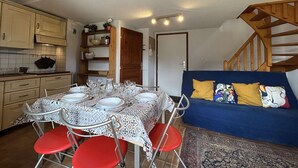 Un espacio habitable compacto con comedor que incluye una mesa preparada para cuatro, un sofá azul con almohadones coloridos y una cocina con armarios de madera y salpicadero de azulejos.
