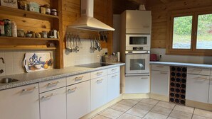 Cocina con armarios blancos, un lavaplatos de acero inoxidable, un microondas y un horno. Hay repisas de madera sobre el lavaplatos y una ventana a la derecha.