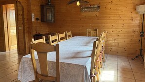Interior de una cabaña de madera con una larga mesa de comedor, sillas, una lámpara colgante y un armario.