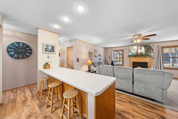A modern kitchen with a bar area, wooden stools, a grey sofa, and a fireplace.