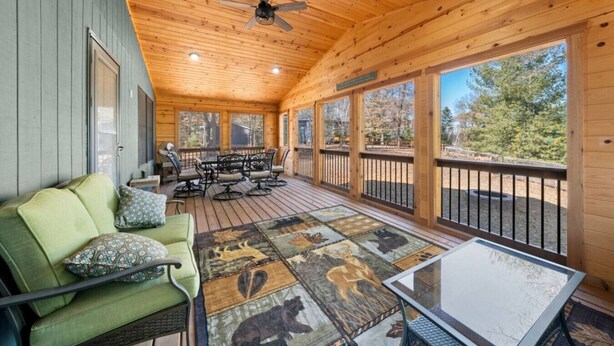 A covered porch with a wooden ceiling, a green sofa, a dining table with chairs, and a view of the outdoors.