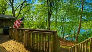 Una terraza de madera con barandilla, una bandera estadounidense y vista a un lago rodeado de árboles.