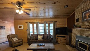 Interior acogedor de una cabaña con techo de madera, chimenea, televisor y un ventanal con cortinas.