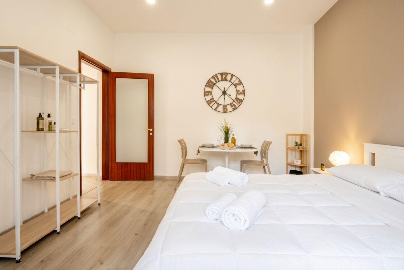 A hotel room with a bed, a wooden wardrobe, a desk with a chair, and a wall clock.