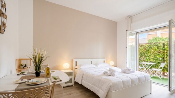 Una camera da letto con un letto, comodini, un tavolino con sedie, una pianta in vaso e vista su uno spazio esterno.