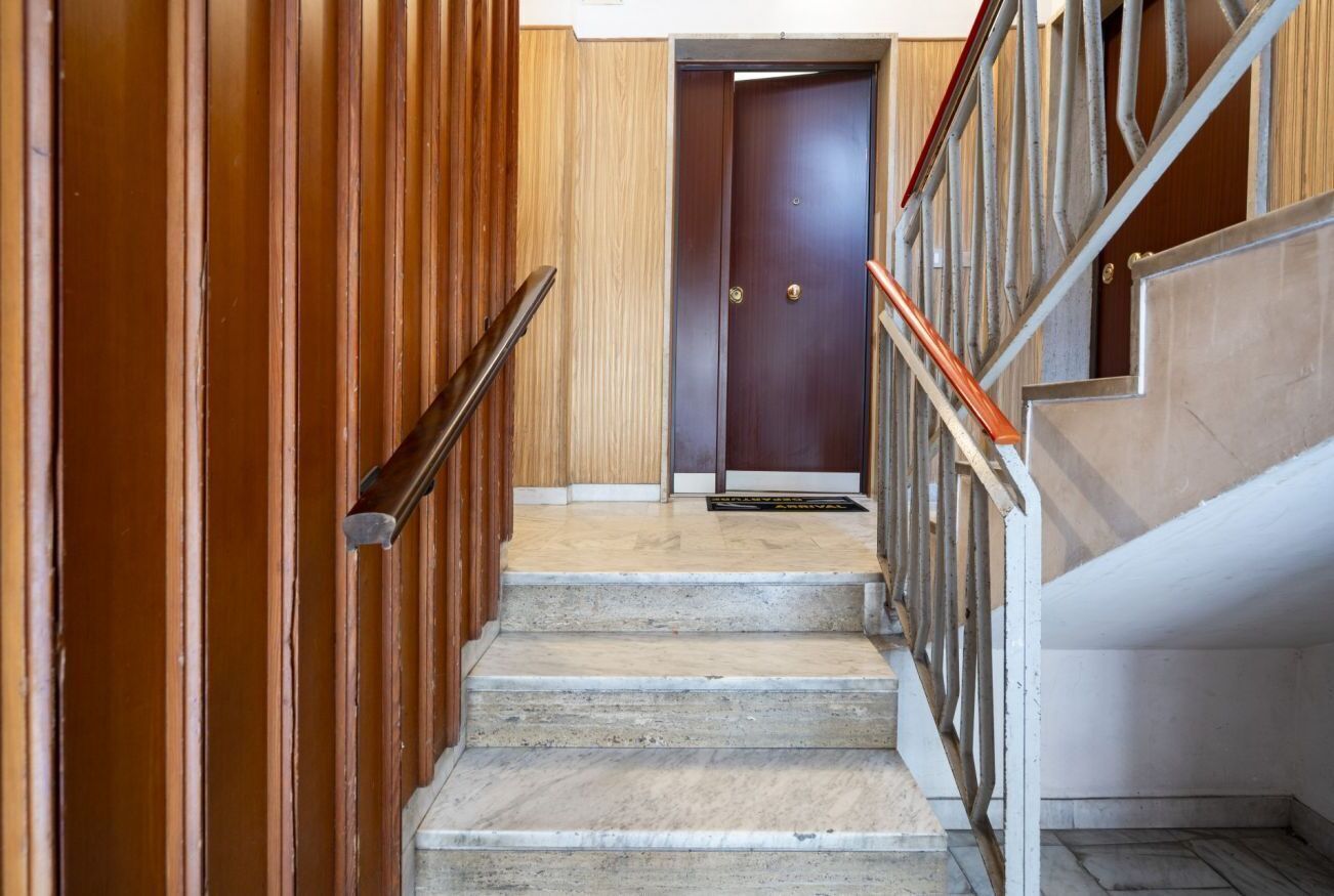 Una scala con rivestimenti in legno su un lato e corrimano in metallo sull'altro, che conduce a un'oscura porta di legno in cima.