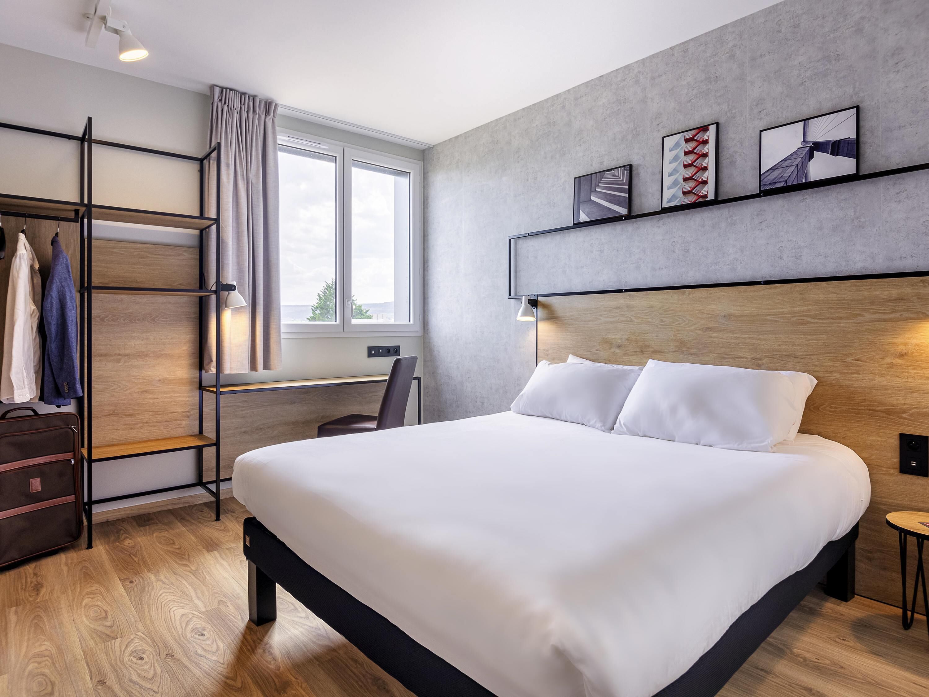A modern hotel room with a large bed, a desk, a wardrobe, and a view of the outdoors.
