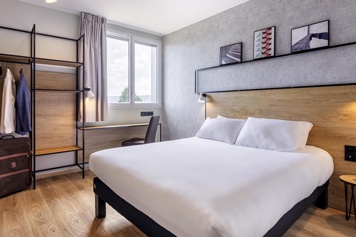 A modern hotel room with a large bed, a desk, a wardrobe, and a view of the outdoors.