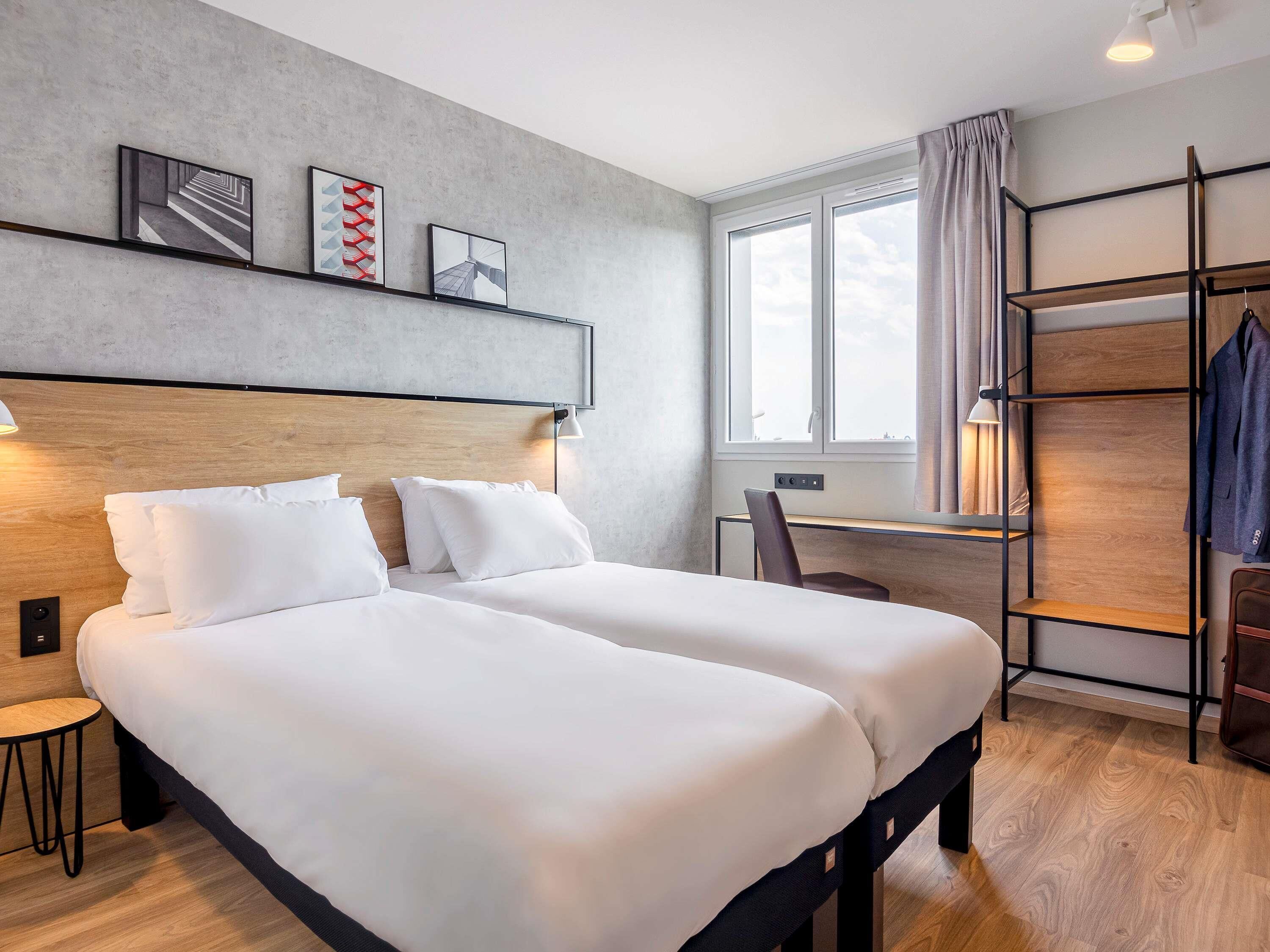 A modern hotel room with two beds, a desk, a chair, a wardrobe, and a window with curtains.