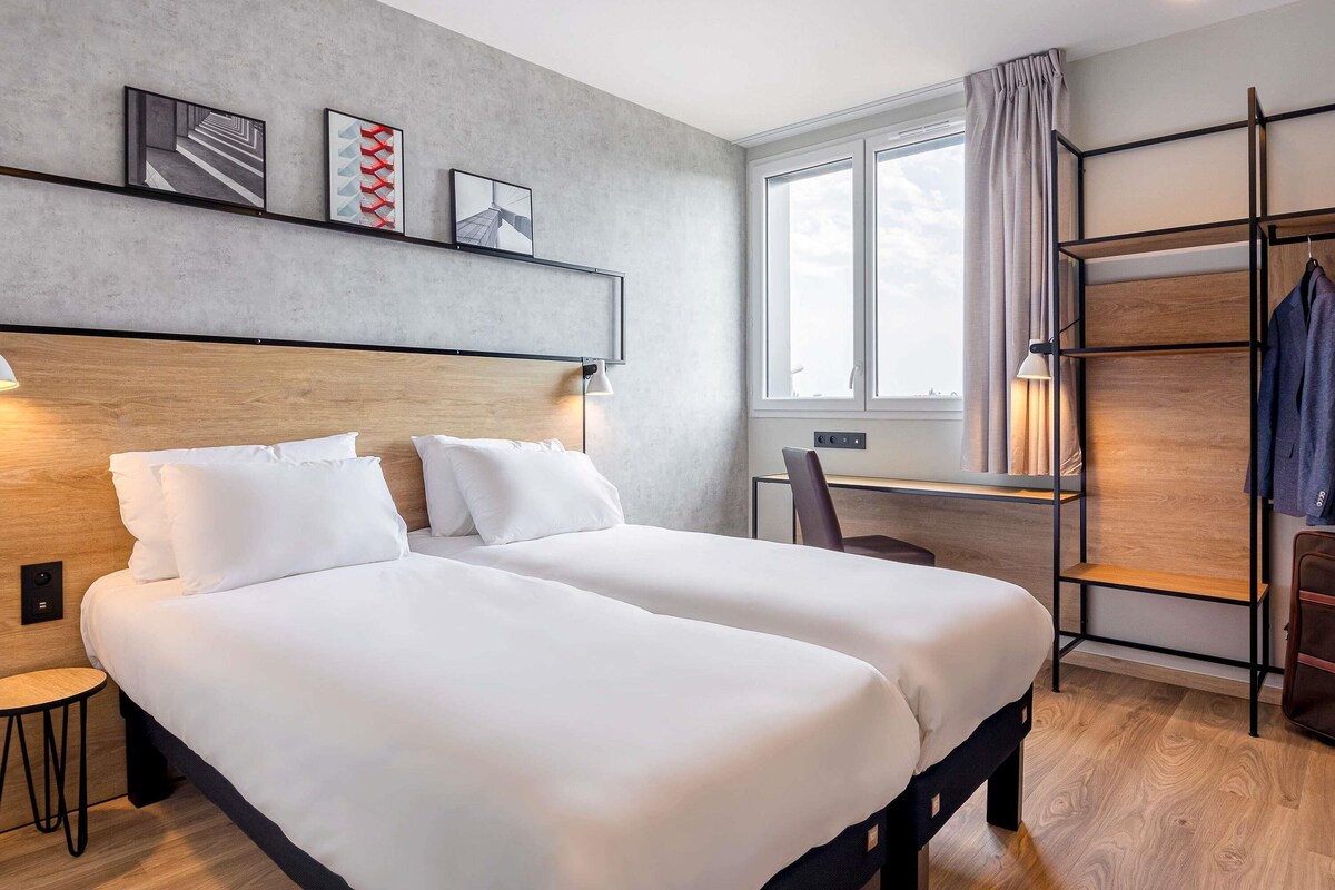A modern hotel room with two beds, a desk, a chair, a wardrobe, and a window with curtains.