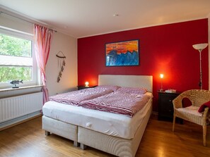 Un dormitorio con una cama, una silla, un cuadro y una ventana con cortinas.