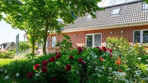 Een bakstenen huis met een pannendak, een tuin vol verschillende bloemen en een boom die schaduw geeft.