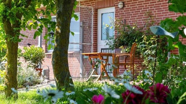 Een bakstenen gebouw met een raam, een houten tafel en stoel, en een boom op de voorgrond.