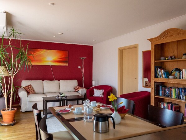Una sala de estar con un sofá, una mesa de comedor dispuesta con tazas y una tetera, una estantería y un cuadro grande en la pared.