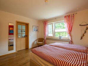 Una habitación con dos camas, una silla, una ventana con cortinas, una puerta y una pared con un cuadro.