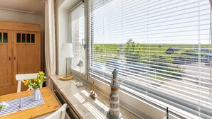 Une pièce avec un meuble en bois, une table ornée d’un vase de fleurs, une lampe, et une vue sur un champ verdoyant par de grandes fenêtres équipées de stores.