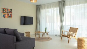A modern living room with a grey sofa, a wooden chair, a TV mounted on the wall, and a large window with sheer curtains. - Alcyone - Gournes NewTown Residences (Heraklion)