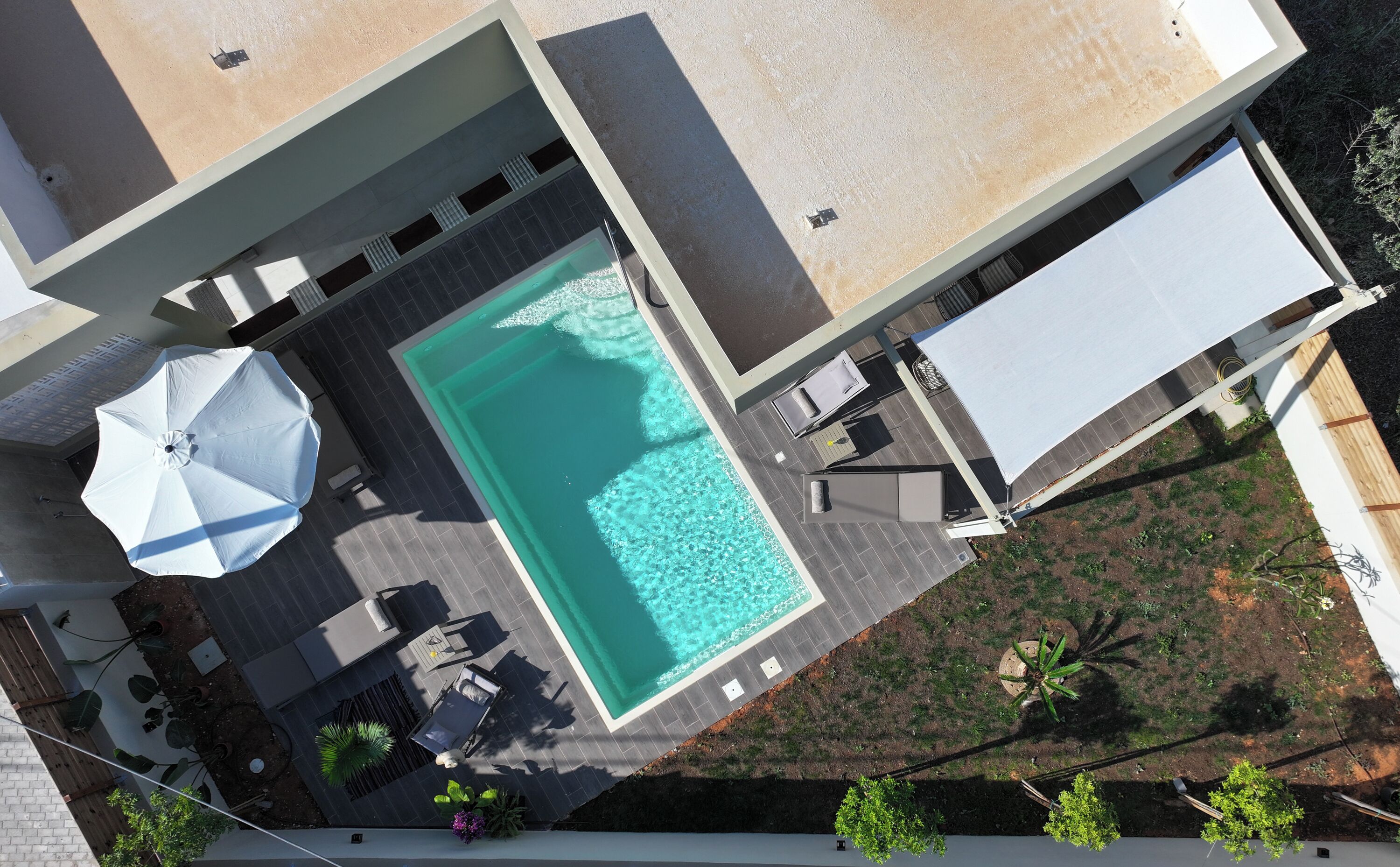 An aerial view of a modern pool area with a rectangular pool, sun loungers, and a covered umbrella.
