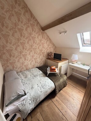 Une chambre confortable située sous les toits, avec un plafond en pente, un lit recouvert d’une literie à motifs, un bureau avec un ordinateur portable et une fenêtre laissant entrer la lumière naturelle.