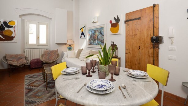A dining area with a round table set for four, surrounded by yellow chairs. There is a potted plant on the table, and the room features a wooden door, white walls, and a window with blinds. - Oltrarno Art House - Florence (Firenze)