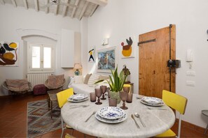 Zona pranzo con un tavolo rotondo apparecchiato per quattro persone, circondato da sedie gialle. Sul tavolo c'è una pianta in vaso, e la stanza presenta una porta in legno, pareti bianche e una finestra con persiane.