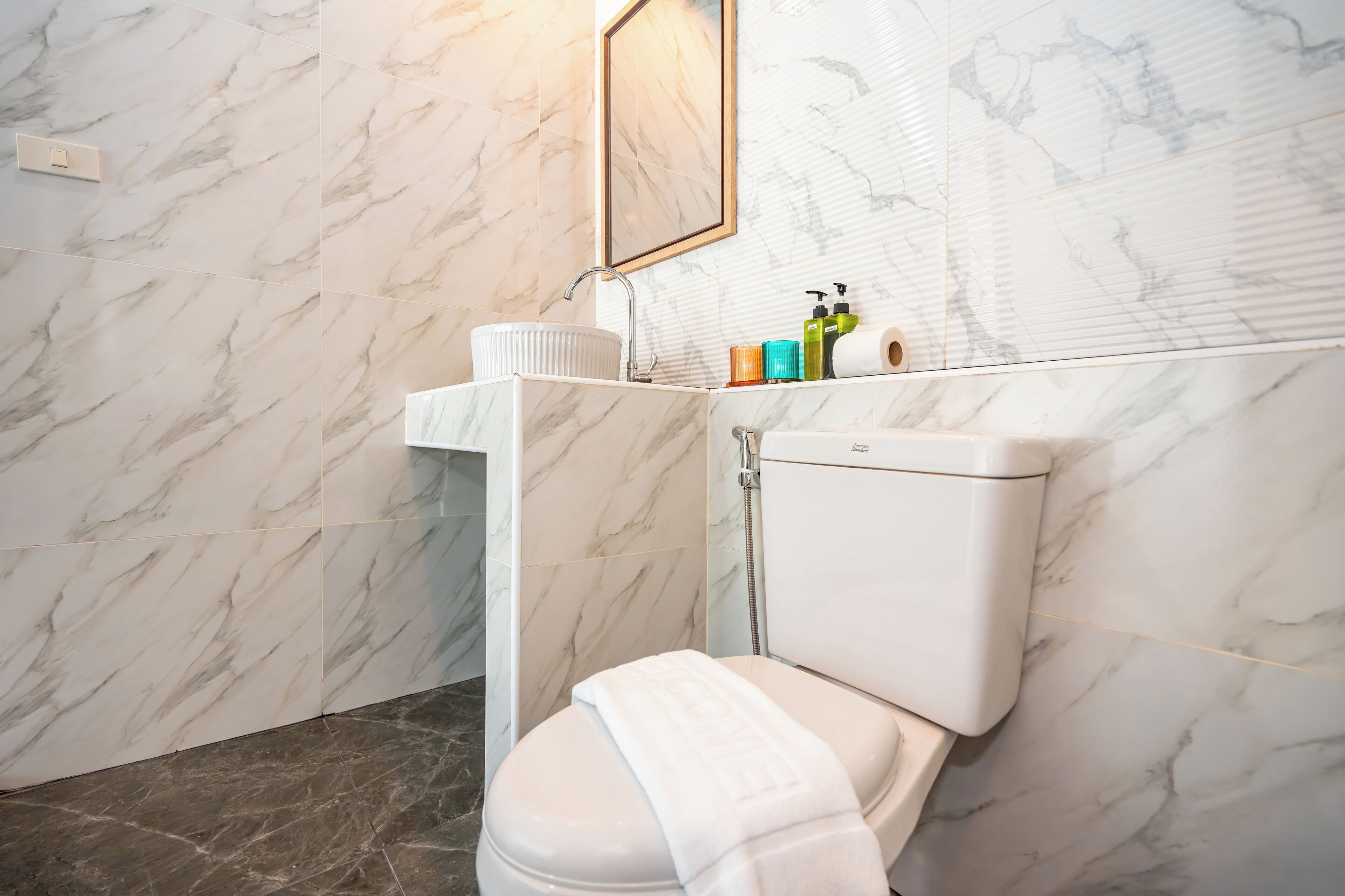 A modern bathroom with a marble wall, a white toilet, a sink with a mirror, and toiletries on the counter.