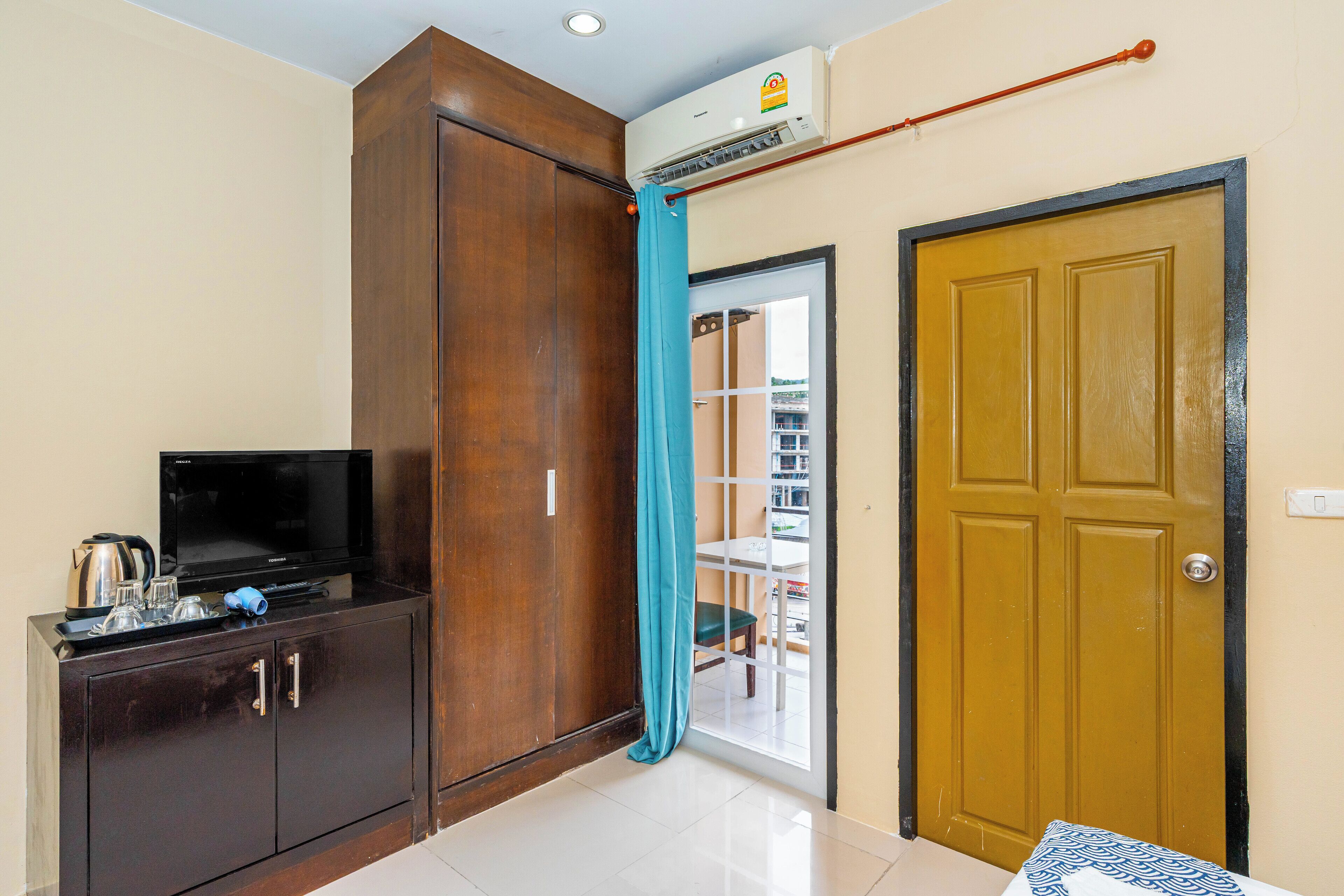 A hotel room with a wooden wardrobe, a television on a cabinet, a door with a curtain, and an air conditioning unit.