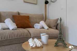 A living room with a beige sofa, a small wooden table with a model of the Eiffel Tower, a white mug, and a plate of whipped cream. - MBA - Diderot 3 - Splendid Apartment (Montreuil)