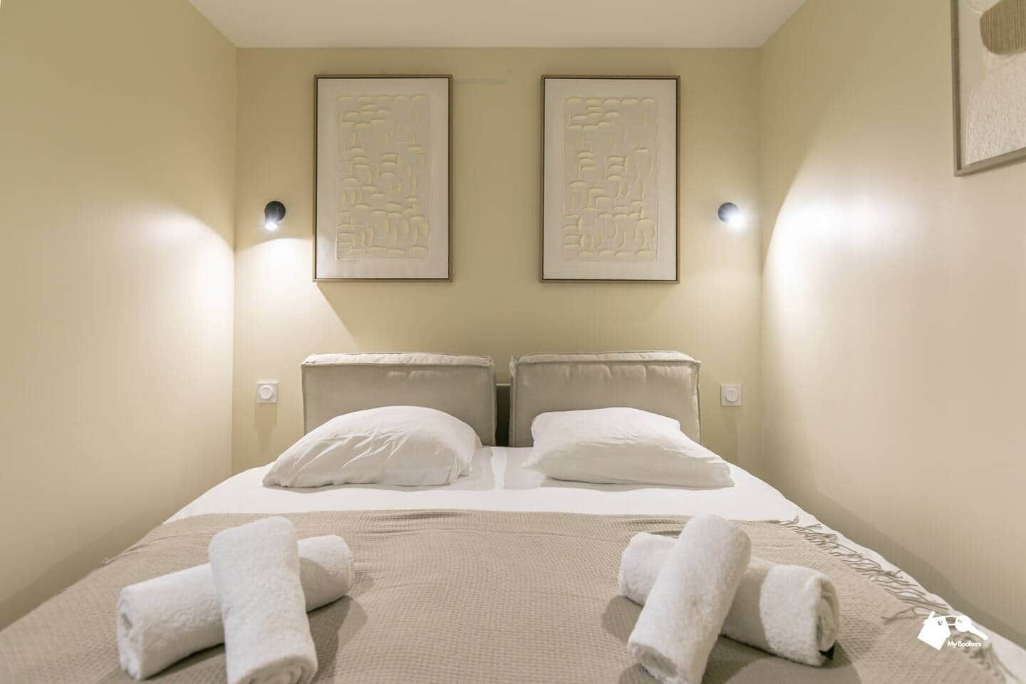 A hotel room with a bed, two headboards, two wall-mounted light fixtures, and two framed wall art pieces.