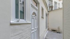 Un estrecho callejón con una puerta y una ventana blancos, una pared de ladrillo y una pequeña zona de concreto con un desagüe.