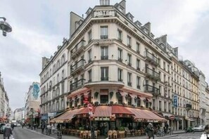 Un edificio en esquina con un letrero que dice 'HOTEL', con varios pisos, balcones y un café en la planta baja con mesas al aire libre.