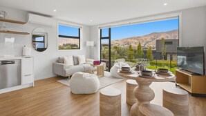 A modern living room with a kitchen, large windows, a flat-screen TV, and a dining area with a mountain view. - Kirimoko Heights (Wānaka)