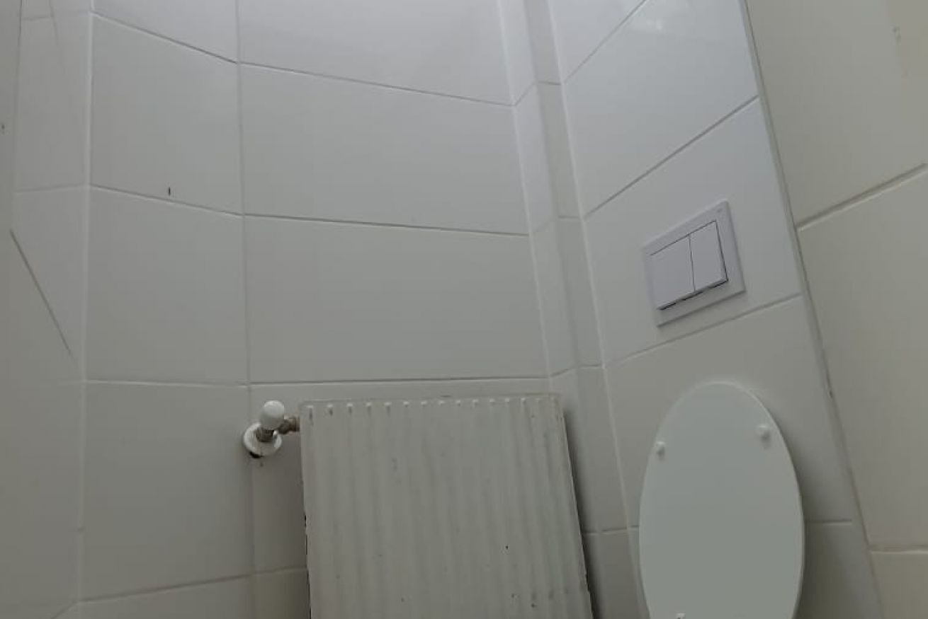 A bathroom with white tiled walls, a toilet, a radiator, and a light switch.