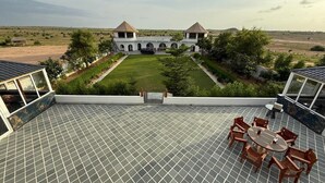 Une terrasse sur le toit, dotée d’un sol carrelé, d’une table ronde entourée de chaises, et offrant une vue sur un bâtiment agrémenté d’un jardin et de structures en arc de cercle.