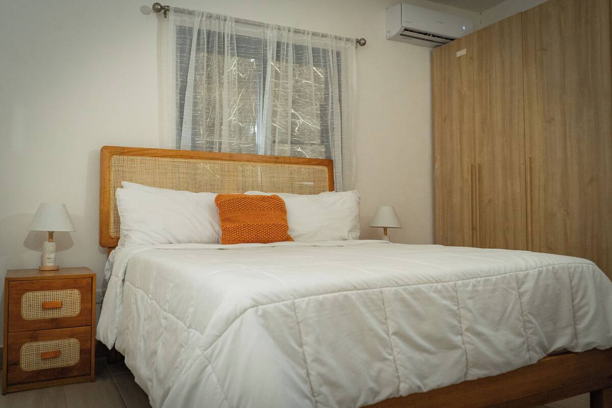 A neatly made bed with white bedding and an orange throw pillow, a wooden nightstand with a lamp, and a large wooden wardrobe.