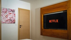 Une chambre avec un lit, une porte en bois, un téléviseur fixé au mur qui affiche le logo « NETFLIX », et un tableau floral accroché au mur.